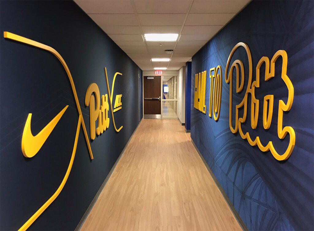 image shows a hallway within a sports facility, likely a locker room or athletic complex, adorned with graphics related to the University of Pittsburgh (Pitt) and the ACC (Atlantic Coast Conference).